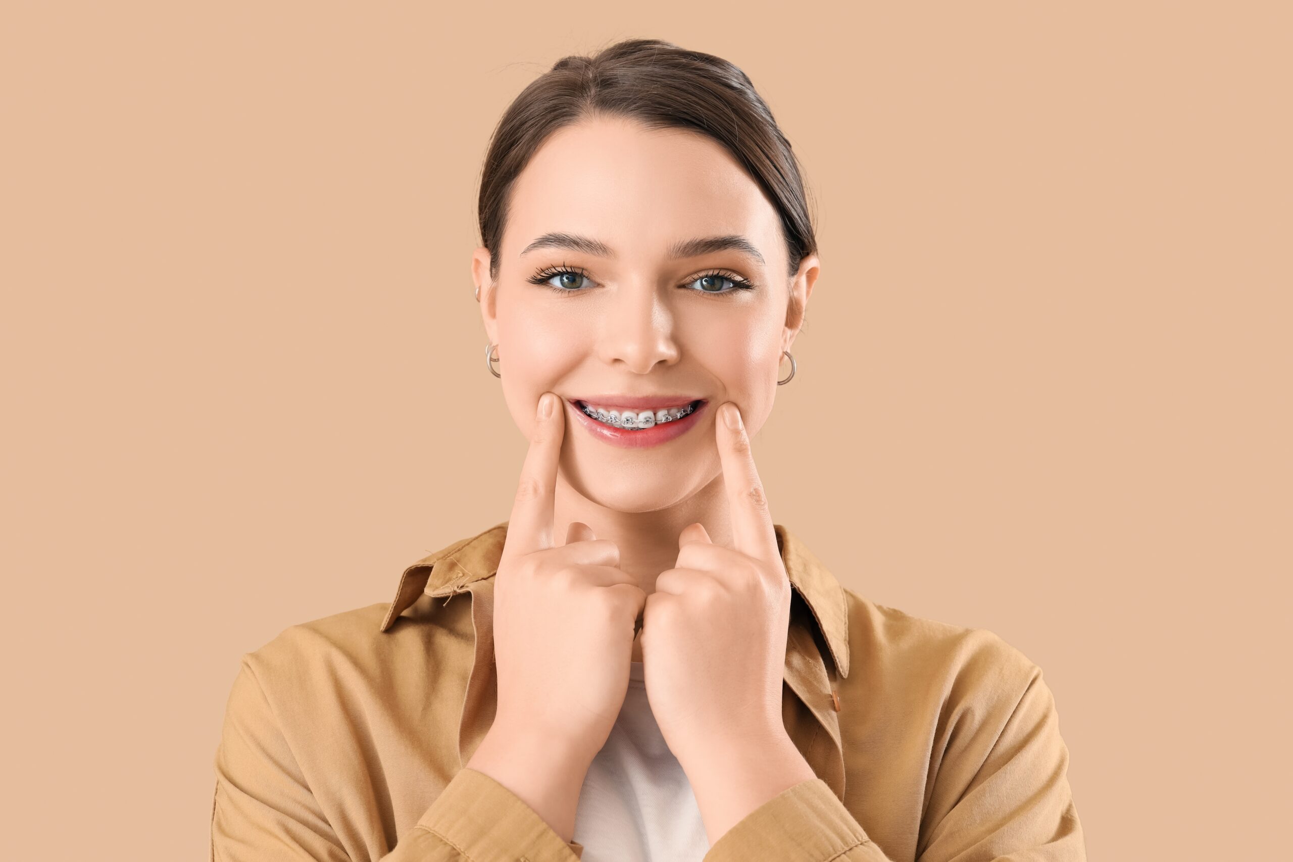 Woman with brown hair and light brown shirt points the corners of her smile to show off straight teeth with braces.