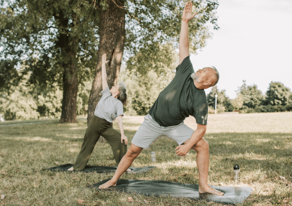 Elder couple seen stretching outdoors in a park as they practice yoga on yoga mats while looking happy and health.