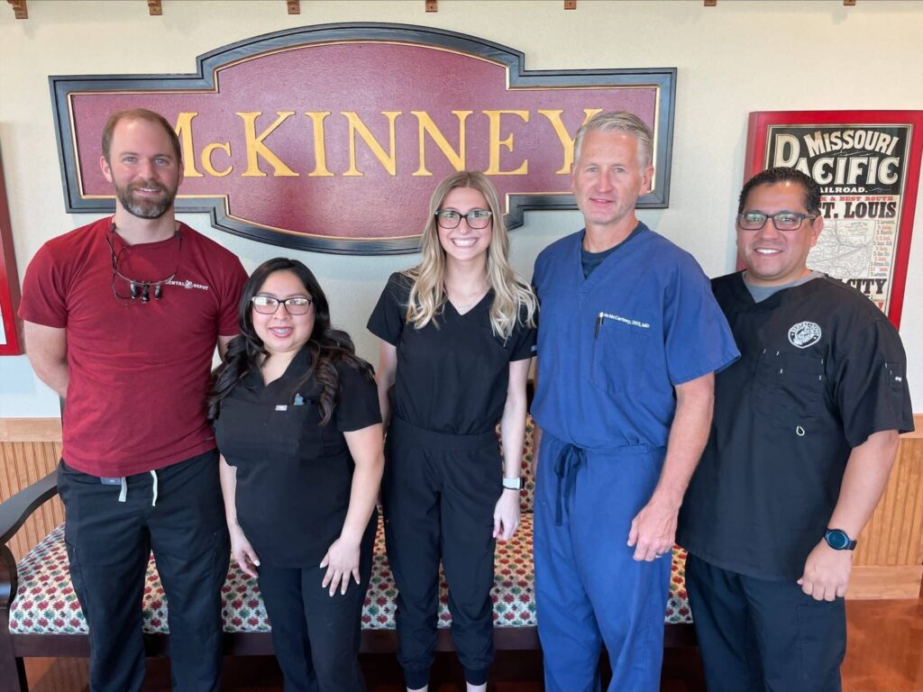 The McKinney dentists and dental team pose for a picture in front of the McKinney train station style logo in the dentist office.