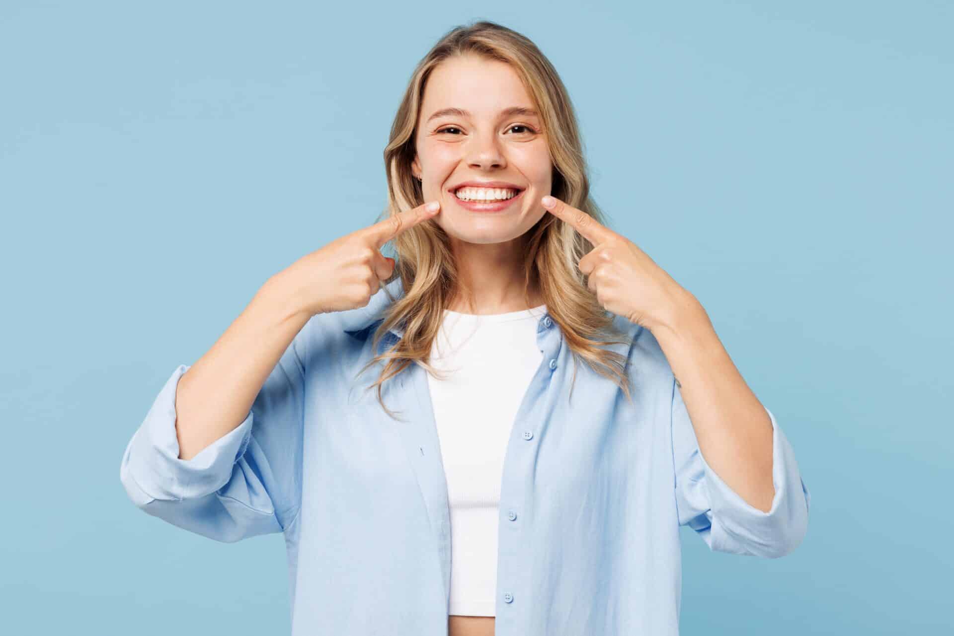 Young woman in casual white top and blue blouse smiles happily while pointing to her bright, white smile against a cyan background.