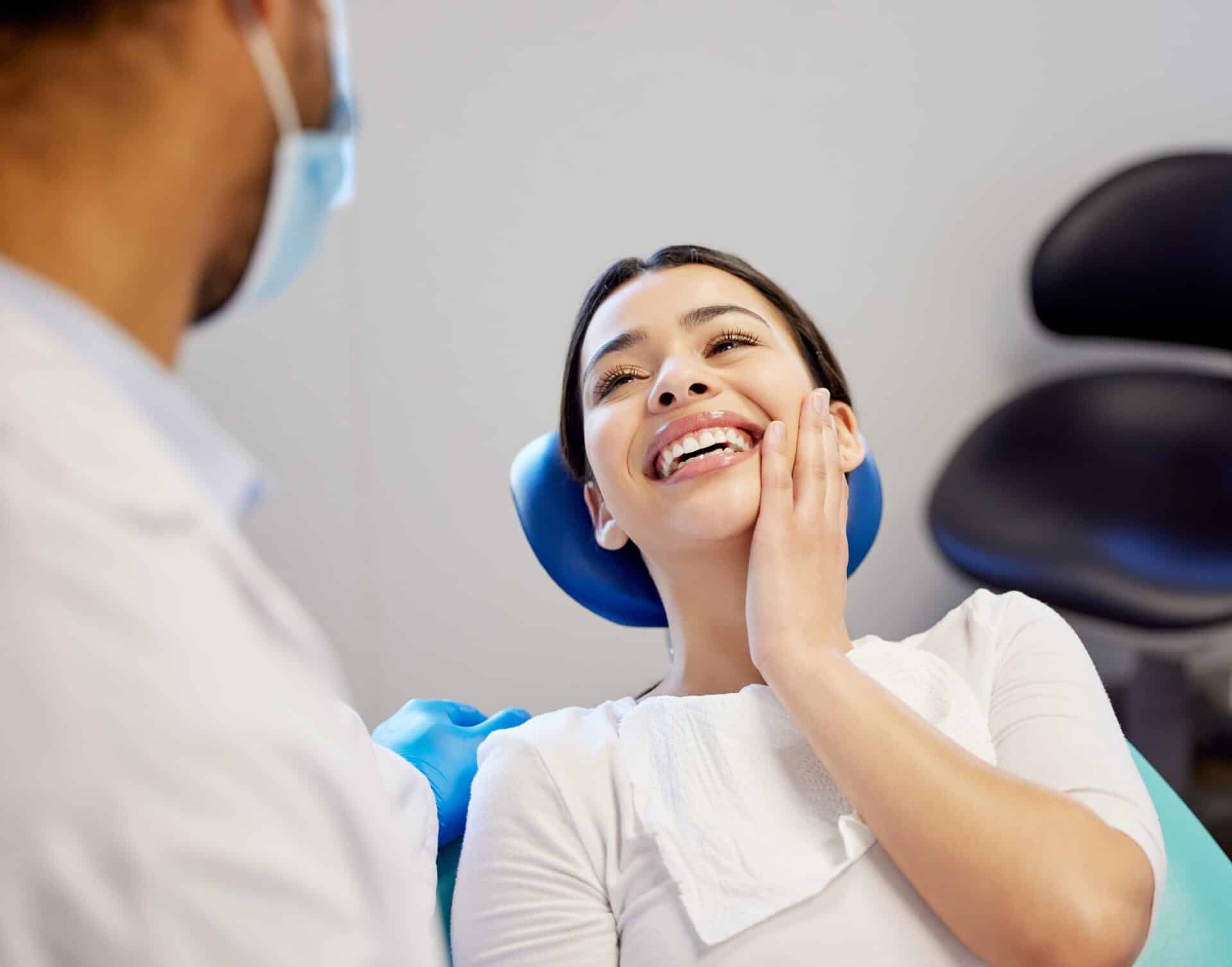 Woman laying in dental chair holds hand to her cheek and smiles at dentist from great oral care.