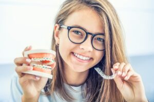 A young woman smiles while holding Invisalign clear aligners and a model of a mouth.